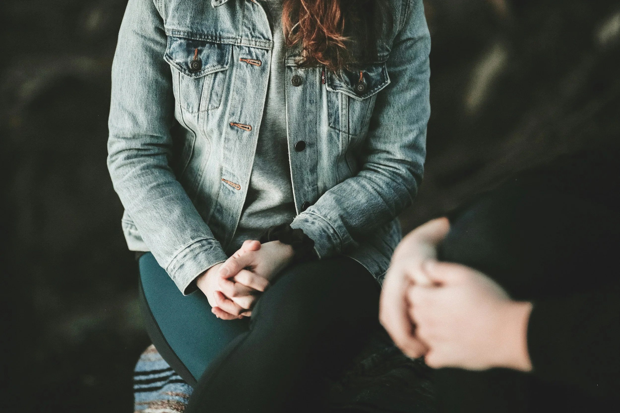 A person wearing a denim jacket and gray shirt seated with hands folded on their lap, engaging in conversation with another person whose hands are visible in the foreground.