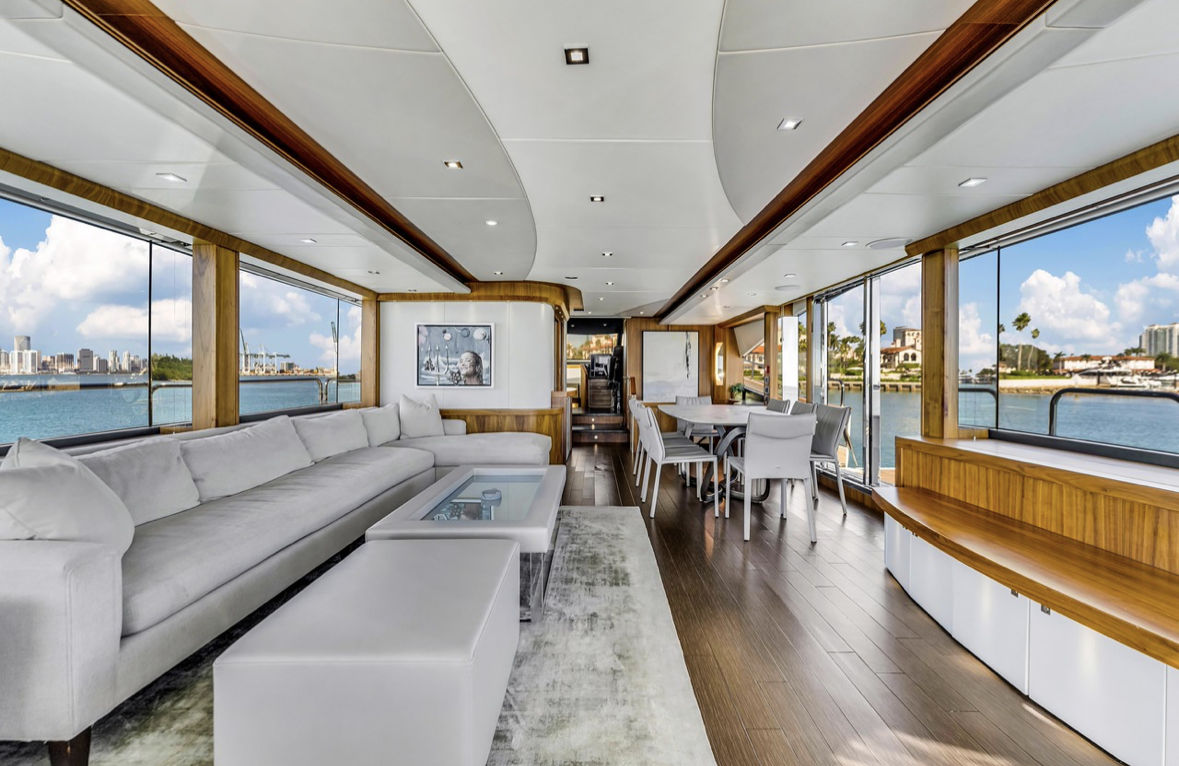 Interior of a yacht with a large white sectional sofa, a dining table with chairs, large windows showing water and city skyline, and wooden accents.