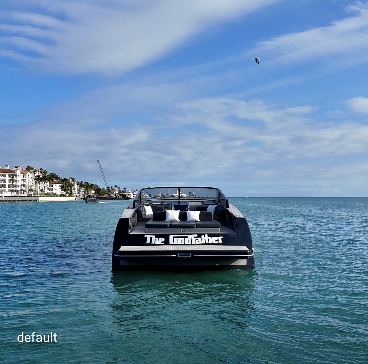 A boat on the water with the words "The Godfather" written on the back, near a shoreline with white buildings and palm trees, under a partly cloudy sky.