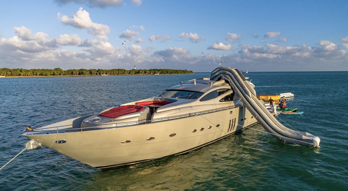Luxury yacht on the water with inflatable water slide and people on small boats nearby, under a partly cloudy sky.