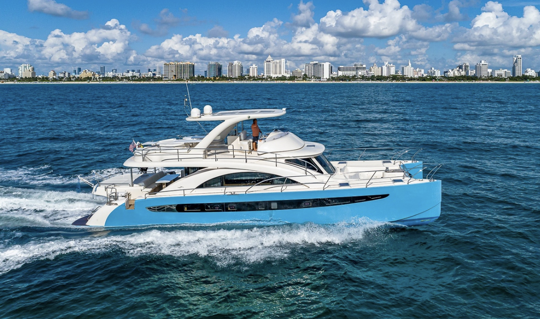 A white and blue yacht sailing in the ocean with a city skyline and partly cloudy sky in the background.