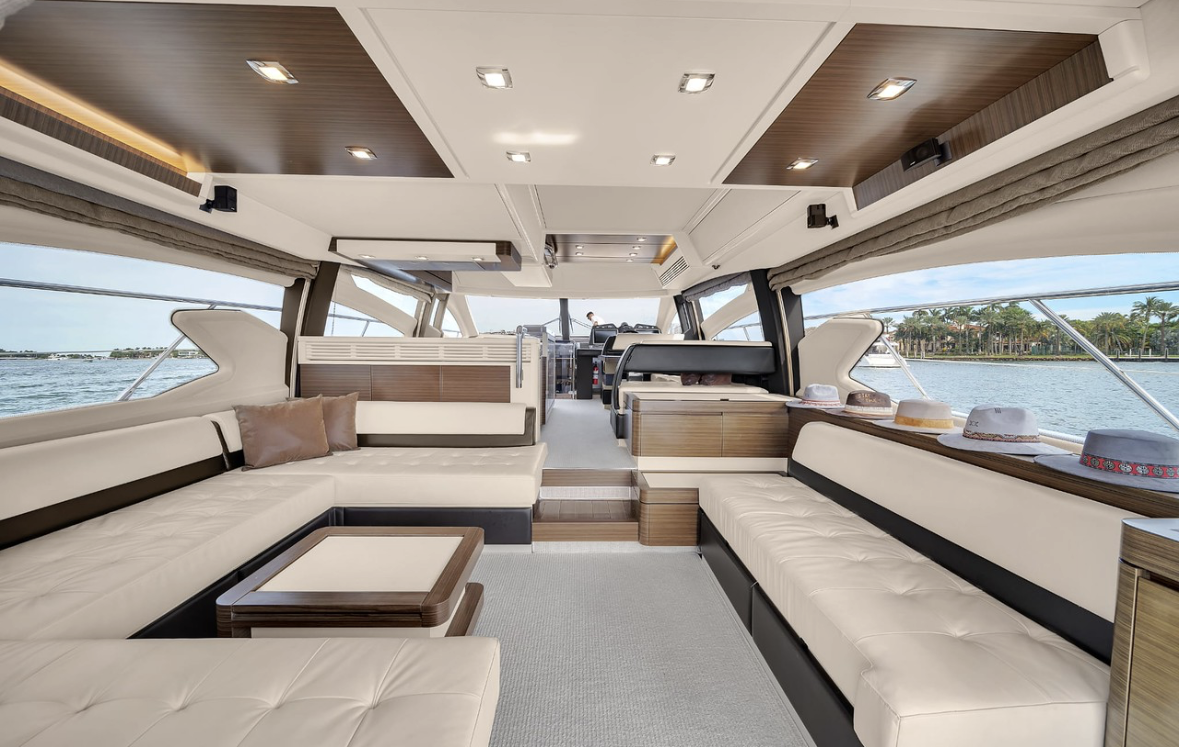Interior of a luxury yacht with cream-colored cushioned seating, a small table, and windows showing water and a shoreline with palm trees outside.