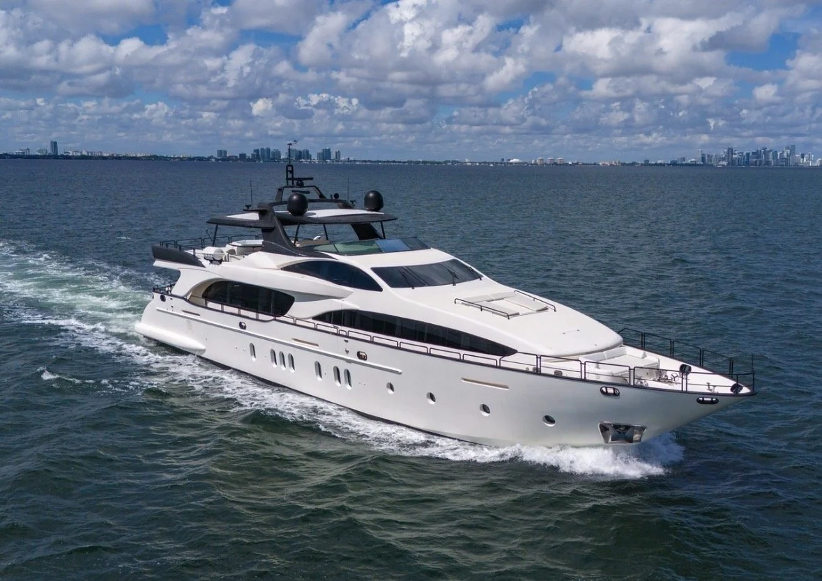 A large white yacht sailing on the water with a city skyline in the background under a partly cloudy sky.