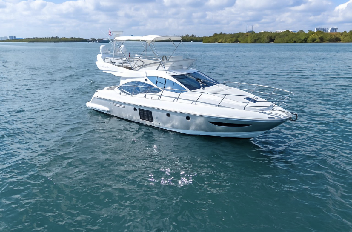 A white yacht floating on a calm body of water with a green shoreline and buildings in the background.