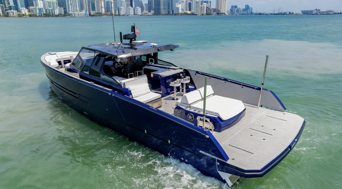 A modern luxury yacht with a sleek dark blue exterior, white seating, and a city skyline in the background, sailing on calm waters.