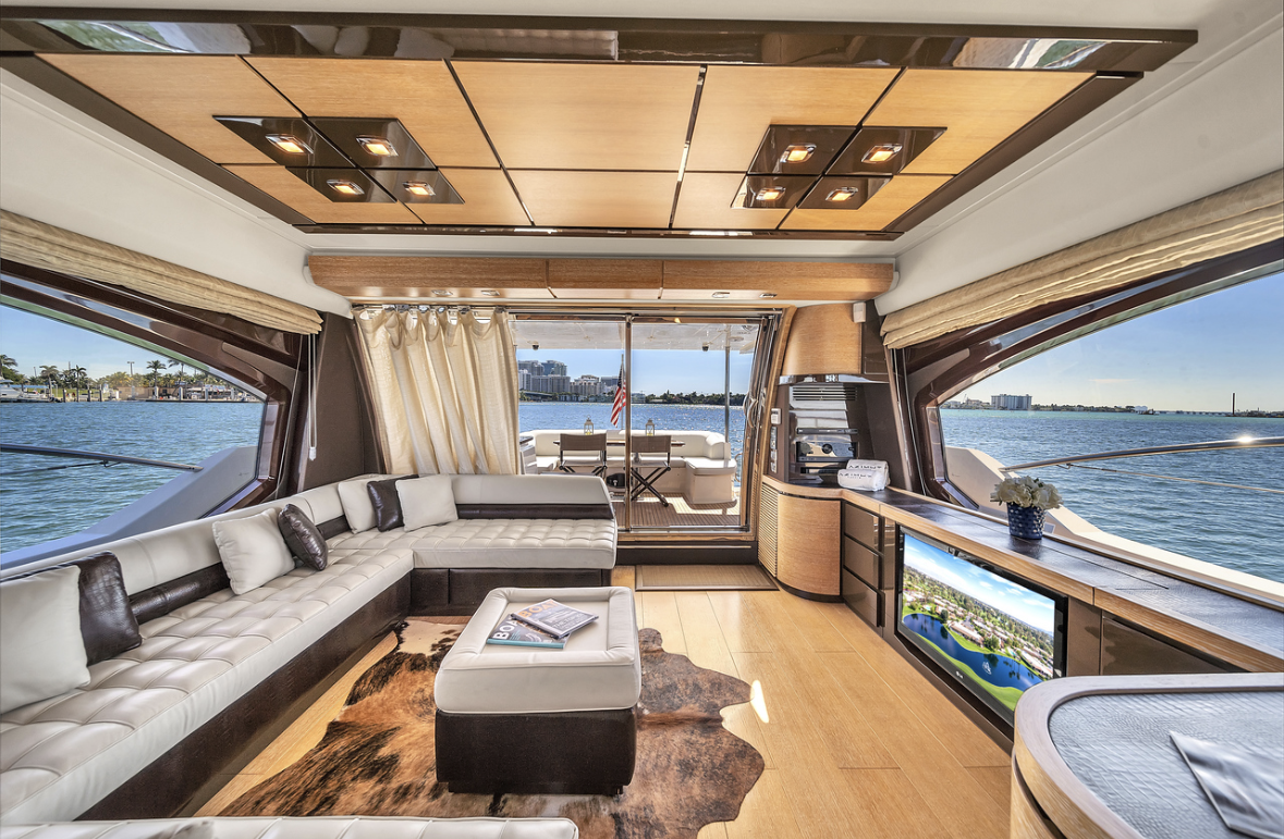 Interior of a yacht with a cream leather sectional, a matching ottoman, a cowhide rug, a wooden ceiling, large windows showing water and city skyline, and a sliding door leading to an outdoor deck with seating and an American flag.