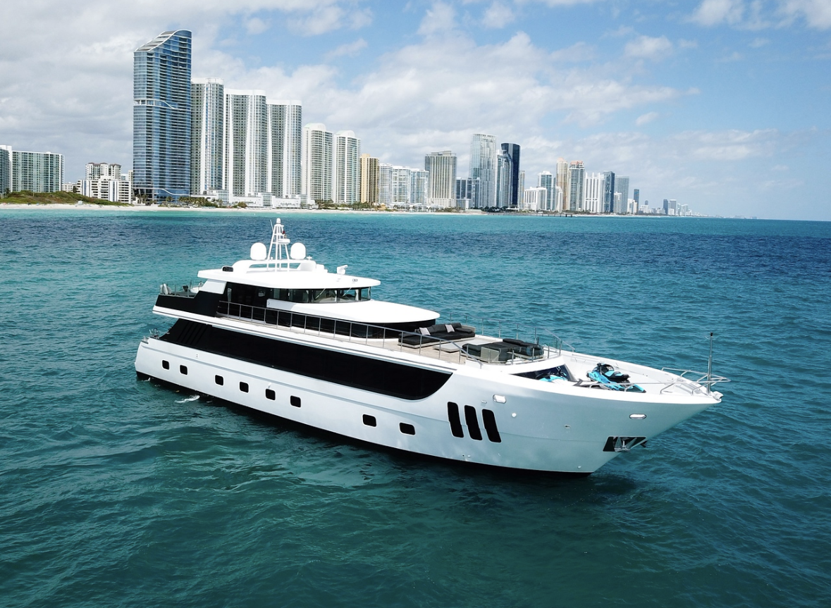 Luxury yacht sailing near a city with high-rise buildings along a coastline, under a partly cloudy sky.