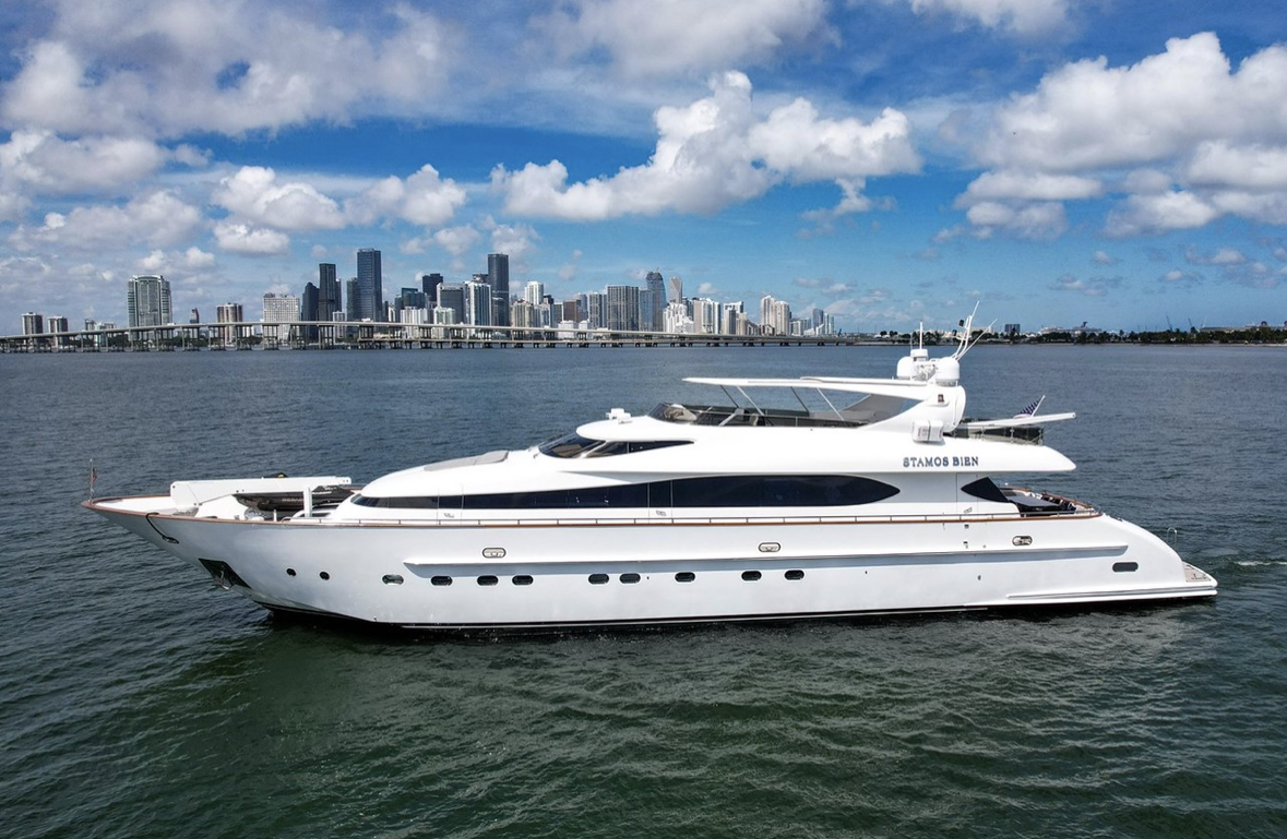 Luxury white yacht floating on water with city skyline and bridge in background, partly cloudy sky.