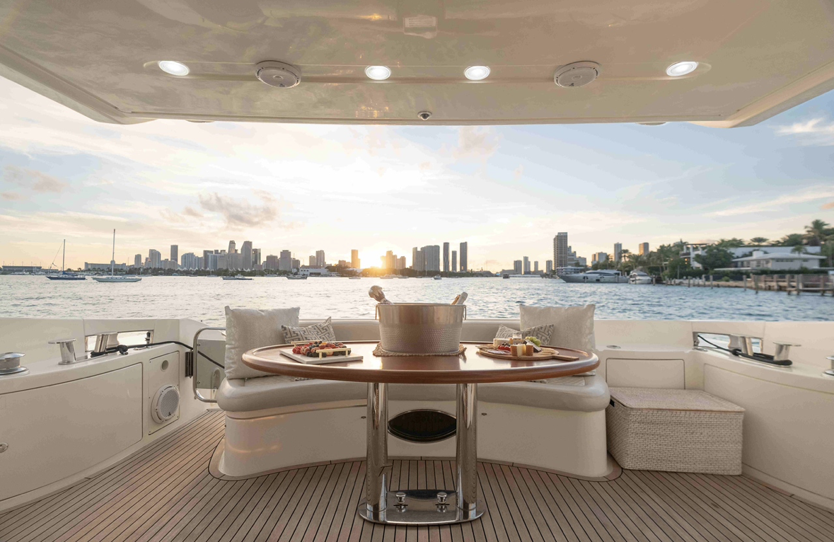 View of a yacht deck with a round table set with food and drinks, overlooking a city skyline at sunset with water and boats in the foreground.