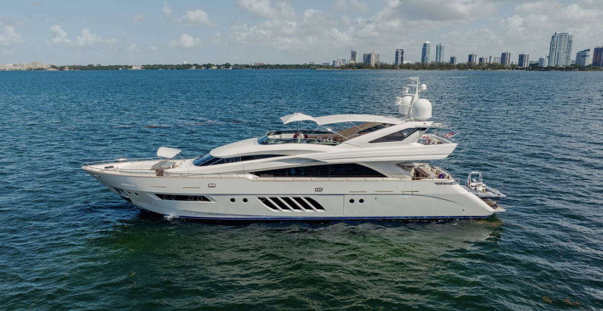 A luxury white yacht sailing in the ocean near a city with high-rise buildings in the background under a partly cloudy sky.