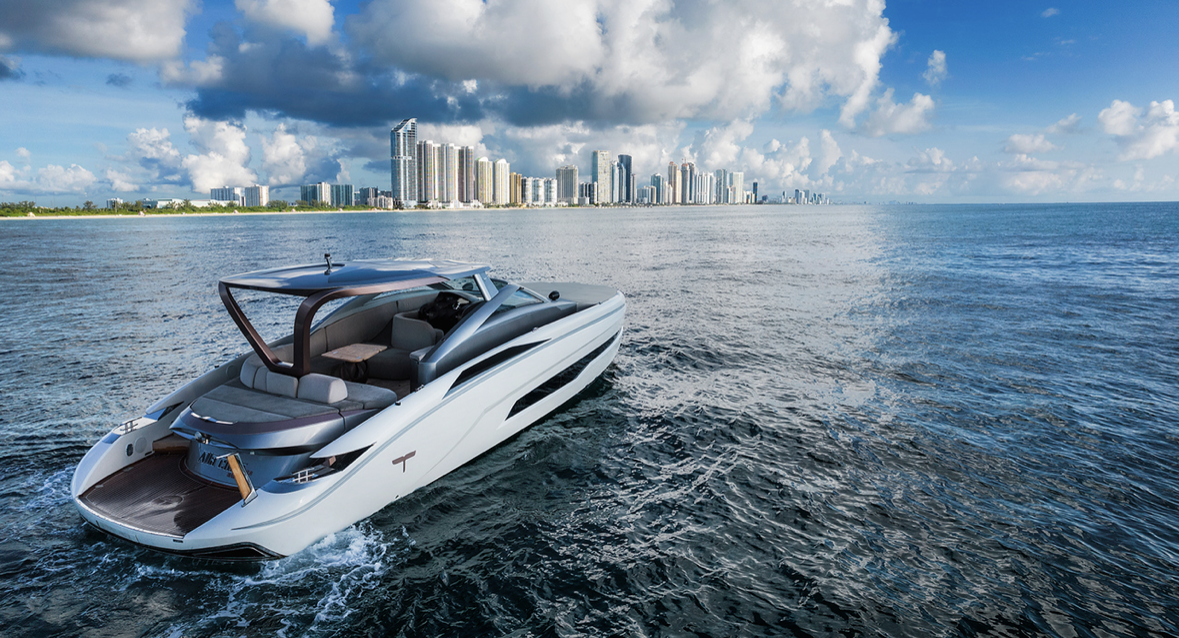A luxury yacht sailing in the ocean with a city skyline in the background and partly cloudy sky.