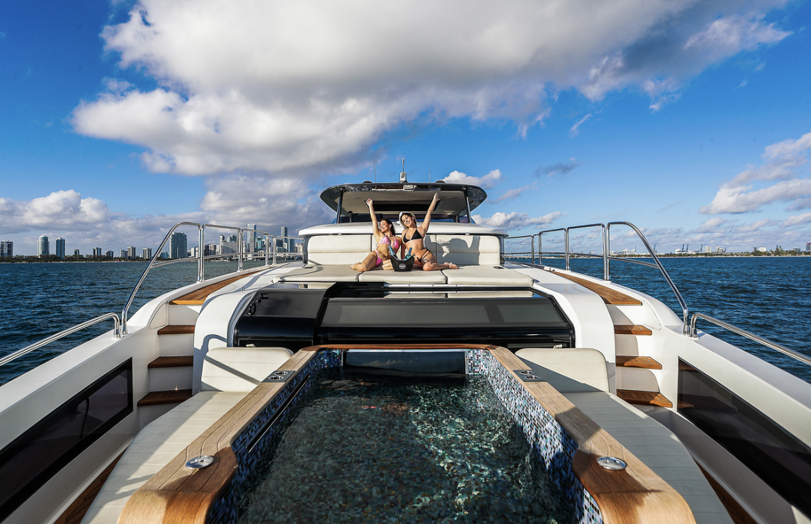 Two women in swimsuits sitting on the sun deck of a luxury yacht, raising their arms in celebration, with a city skyline and water in the background on a partly cloudy day.