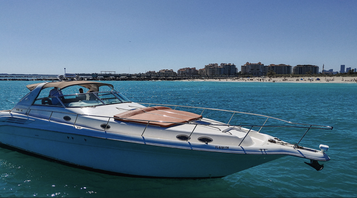A white yacht anchored in a clear blue ocean with a city skyline and sandy beach in the background on a sunny day.