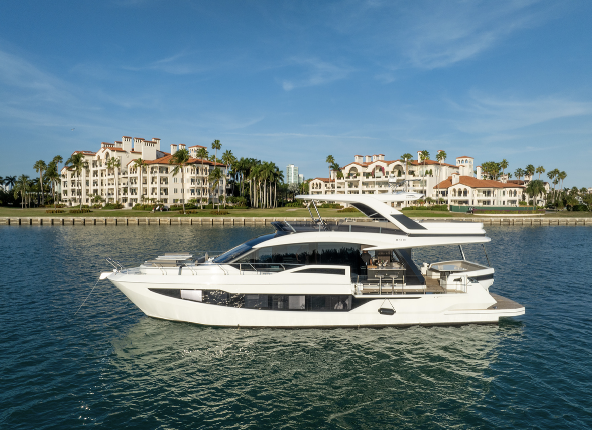 A white yacht sailing on calm water with luxurious residential buildings and palm trees in the background under a blue sky.