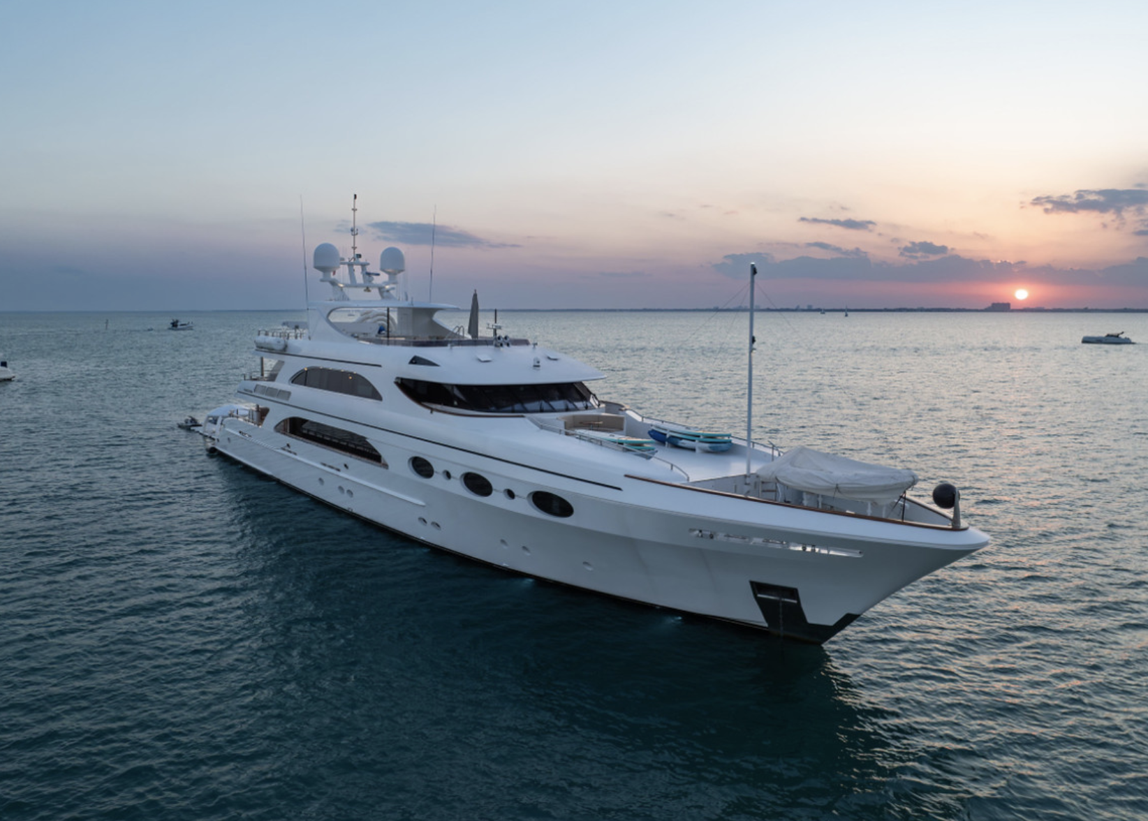 A luxurious white yacht floating on calm water at sunset with several smaller boats in the distance.
