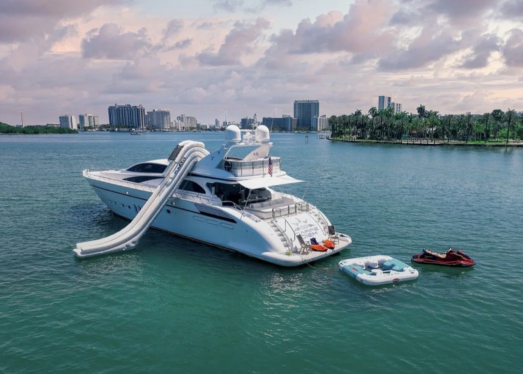 Large white yacht with an inflatable slide on the side and an upper deck, anchored in a bay near a city with high-rise buildings and palm trees in the background. There are two smaller boats and a floating raft nearby.