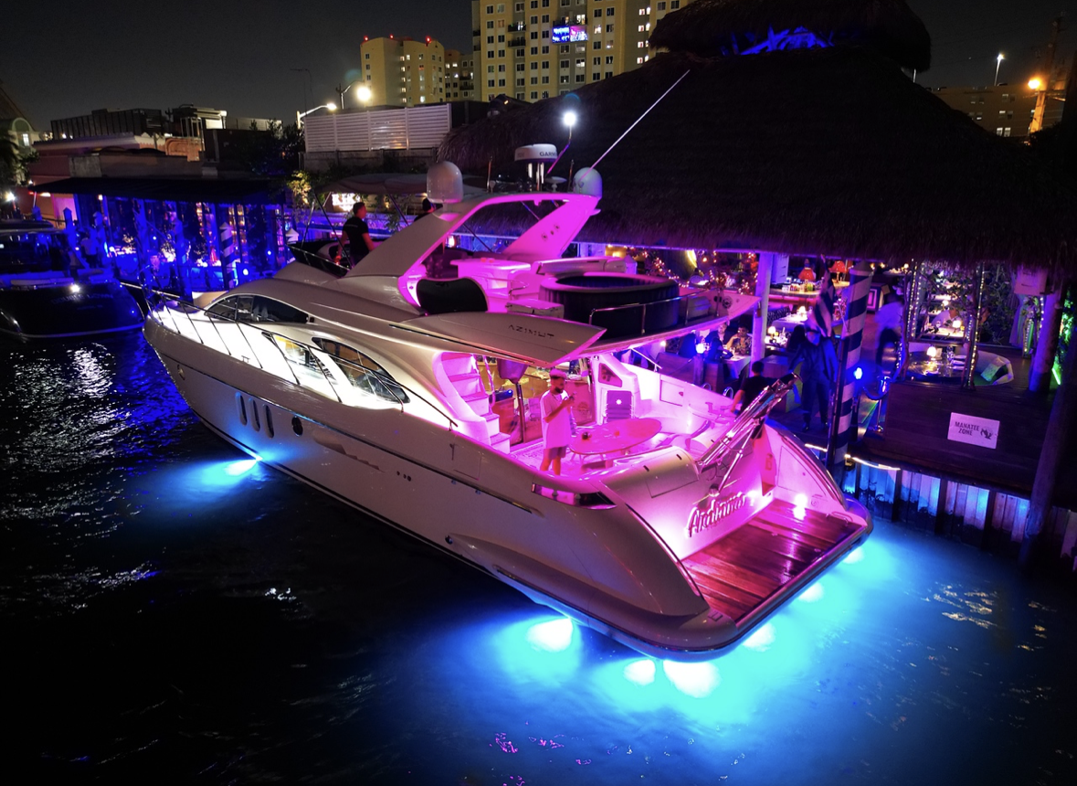Luxury yacht illuminated with pink and blue lights docked at a waterfront at night, with a active bar and restaurant on the dock under a thatched roof.
