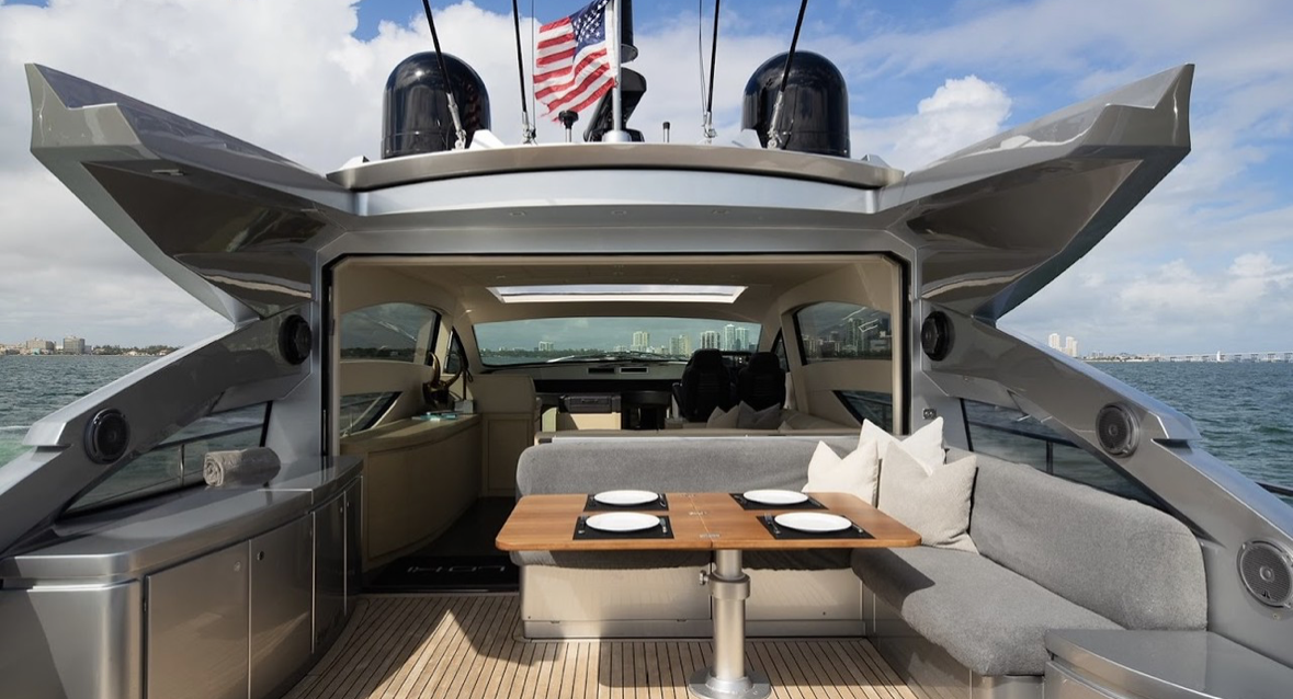 Interior of a luxury yacht with a dining table set with plates, surrounded by cushioned seating, and an open view through the front windows showing water and a city skyline.