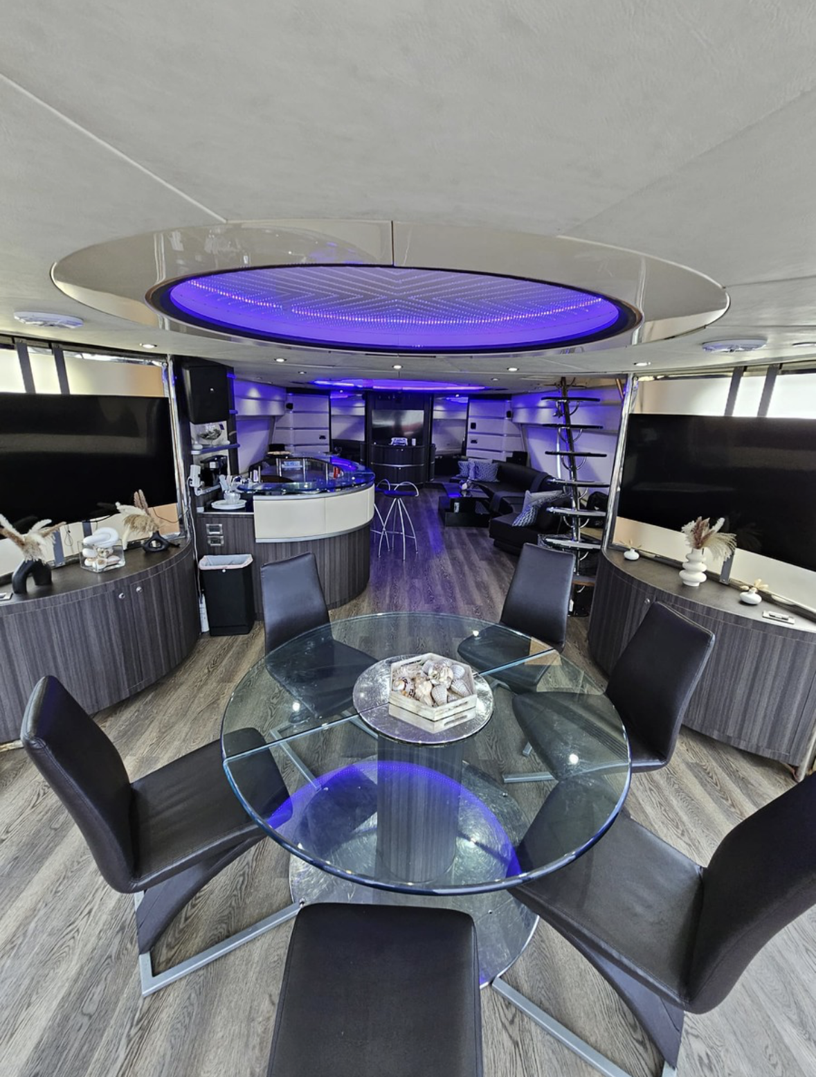 Modern lounge with a round glass dining table, black chairs, a bar area with high stools, and seating area with black sofas and cushions, illuminated with blue and purple lighting.