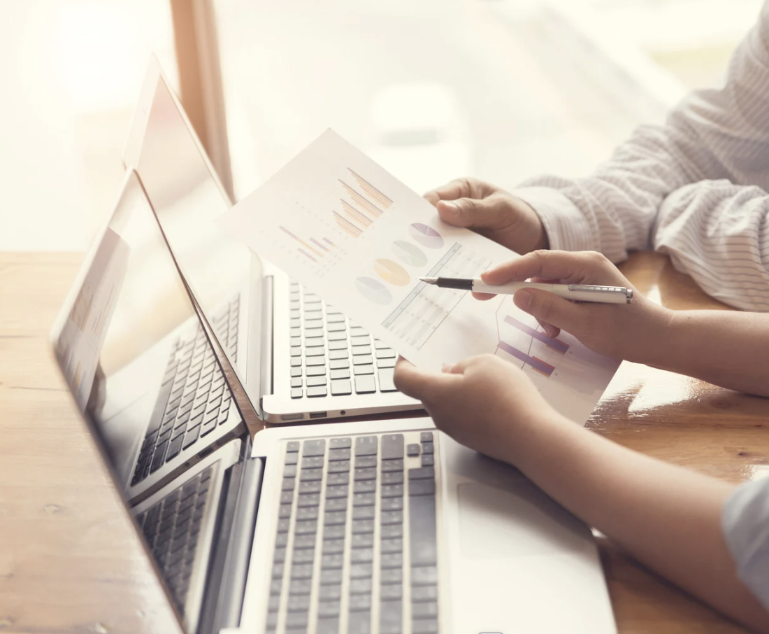 Two people at a wooden desk with open laptops, reviewing printed charts and graphs.
