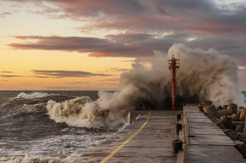 Waves crashing against a breakwater at sunset with a lighthouse and stormy sky.