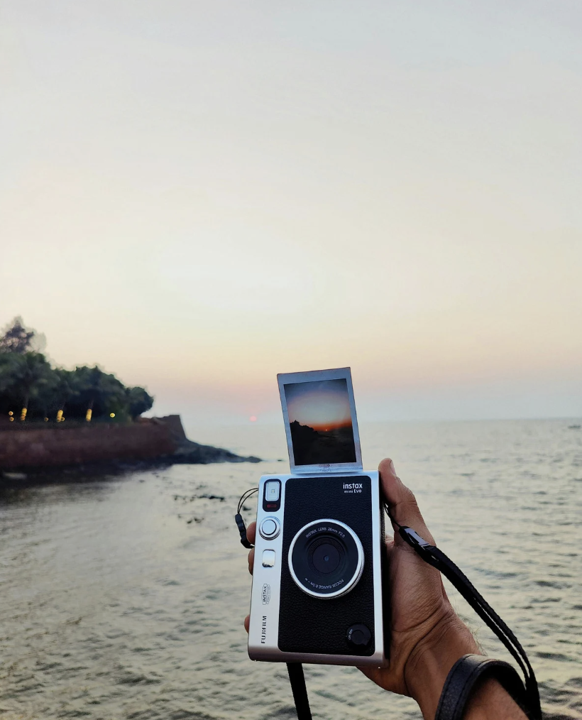 Person holding a vintage Fuji Instax camera capturing a sunset over the ocean with a small island and trees in the background.