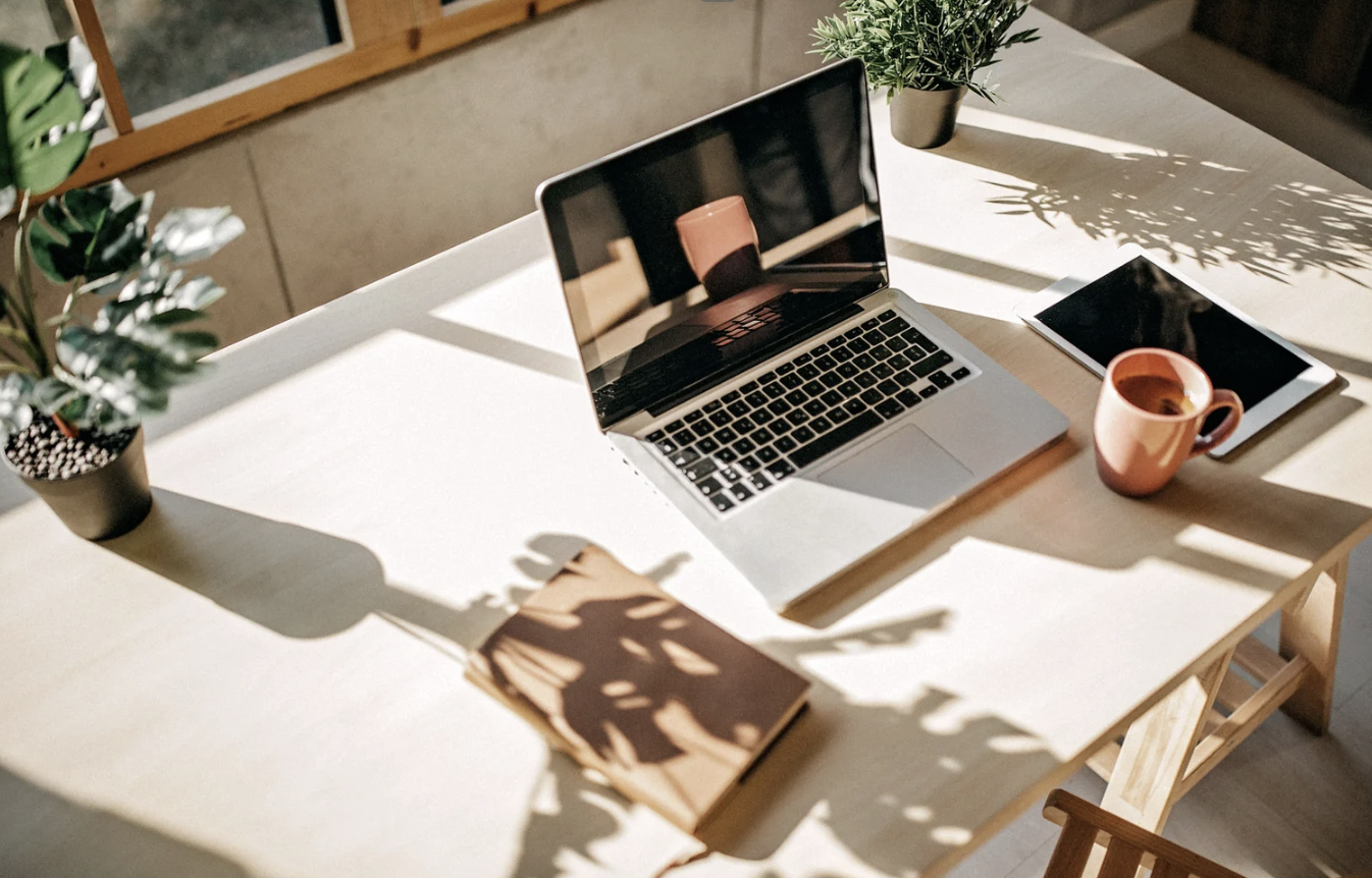 A bright workspace with a laptop, tablet, coffee mug, notebook, and potted plants on a white table, with shadows cast by sunlight.
