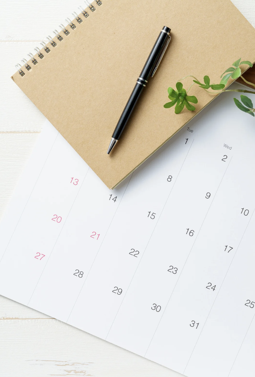 A black pen placed on a spiral notebook with a blank kraft cover, resting on a white desk. Next to it is a partial view of a printed calendar displaying the dates of a month, with some dates in pink and others in black, along with a small green plant leaf.