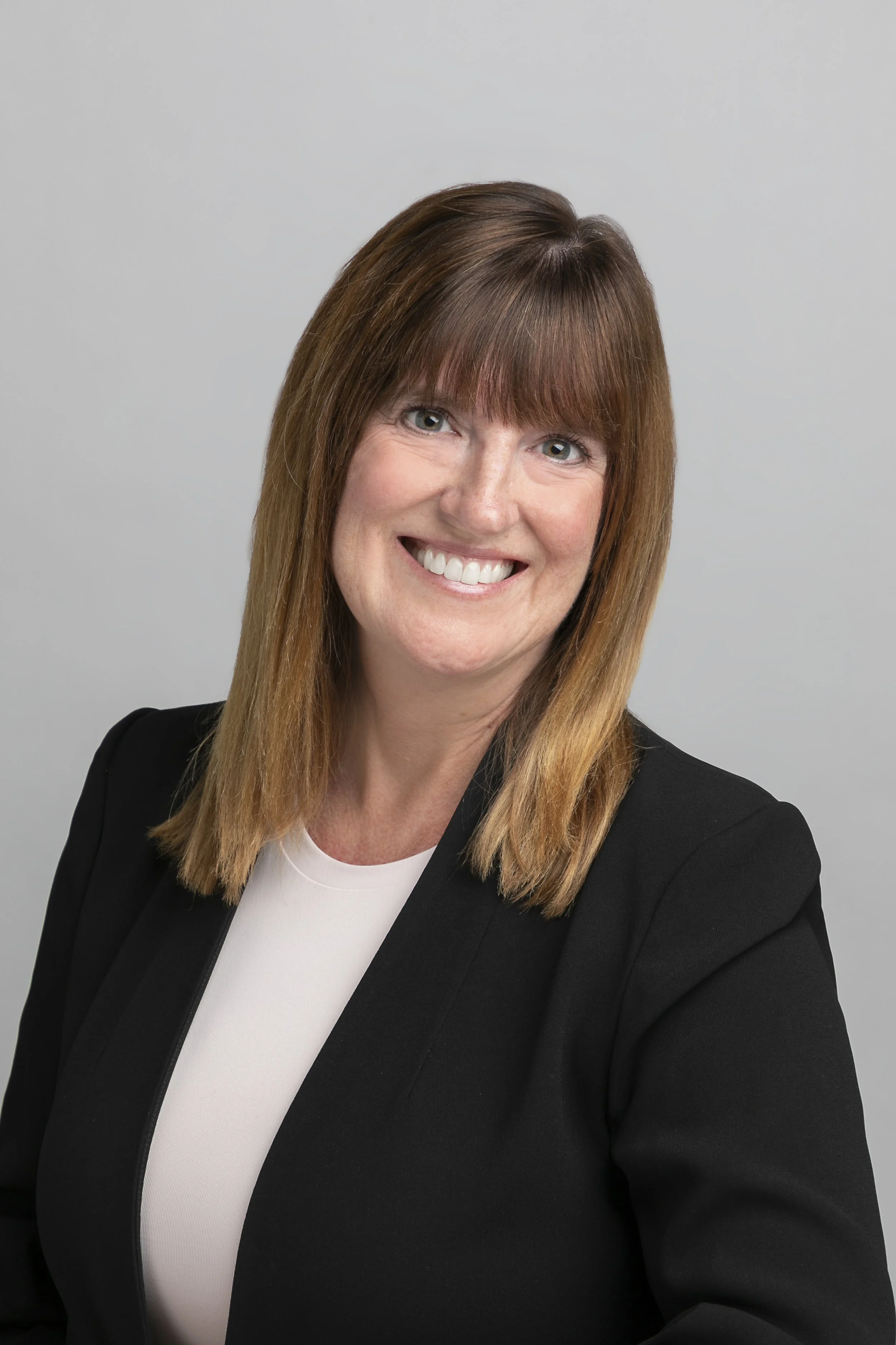 A woman with shoulder-length light brown hair and bangs, smiling in front of a plain light gray background, wearing a black blazer over a white top.