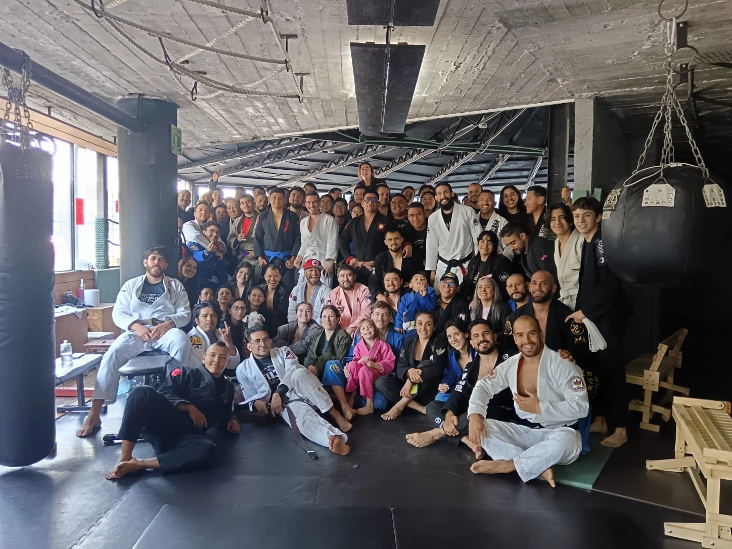 Group photo of Brazilian Jiu-Jitsu practitioners and instructors in a gym, some wearing gis, posing together on mats in front of punching bags.