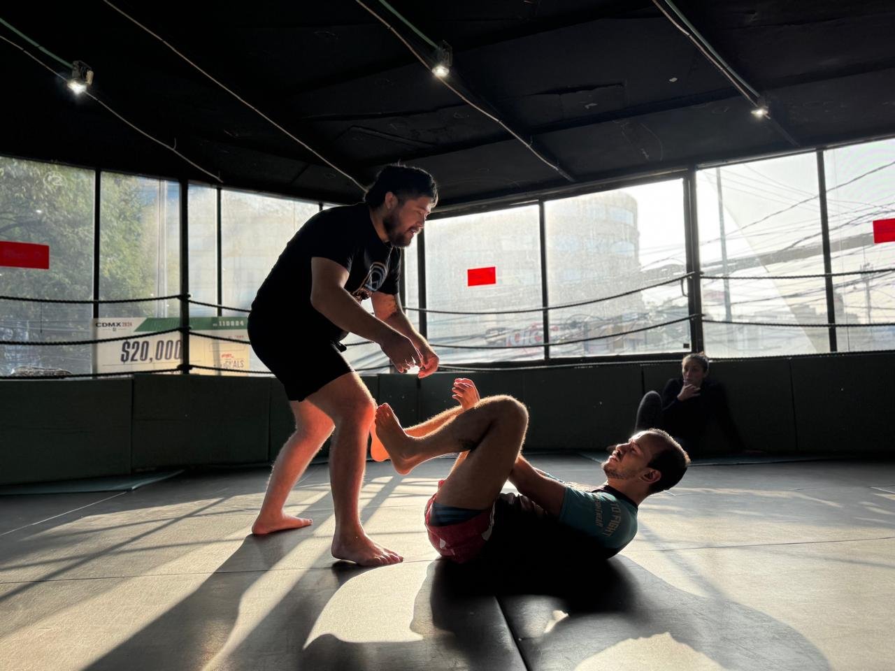 Two men practicing Brazilian Jiu-Jitsu in a training gym, with one man on the ground and the other standing, both smiling, and a woman sitting in the background watching.