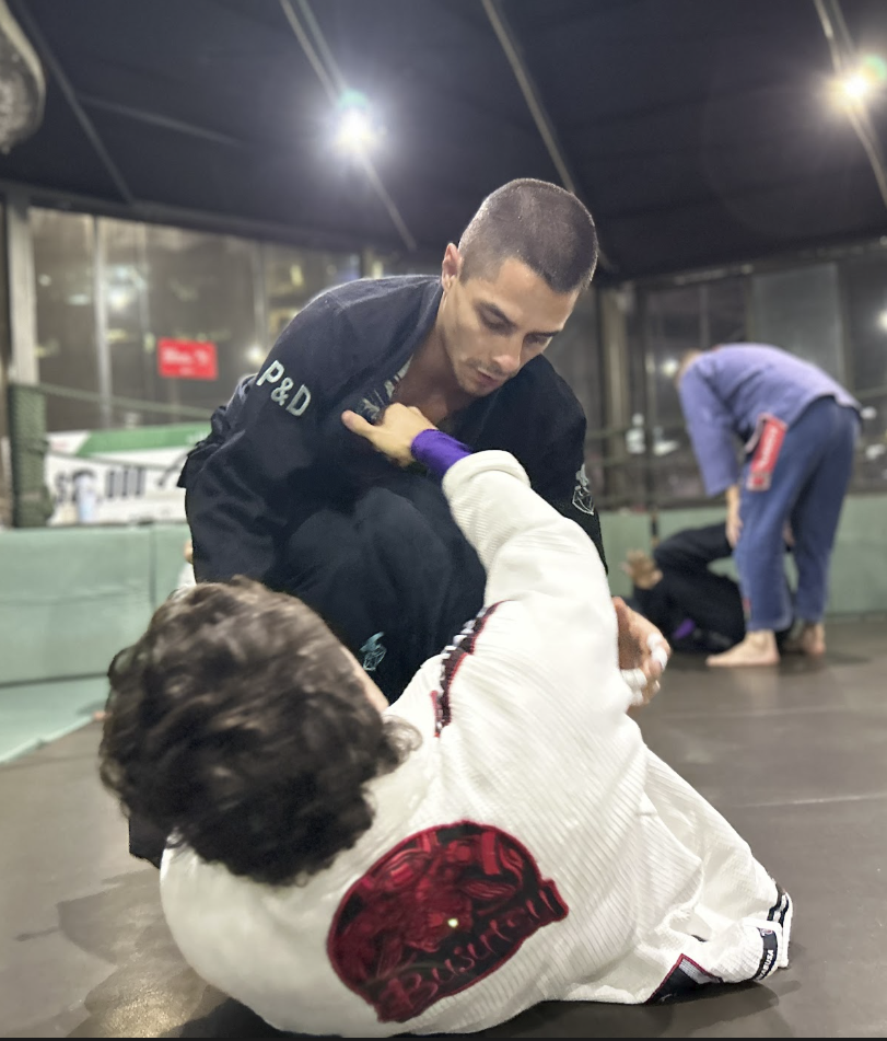 Two people practicing Brazilian Jiu-Jitsu on a mat in an indoor gym, with one person in a black gi and the other in a white gi. The person in the white gi is on the ground attempting a guard position, while the person in the black gi is leaning over.