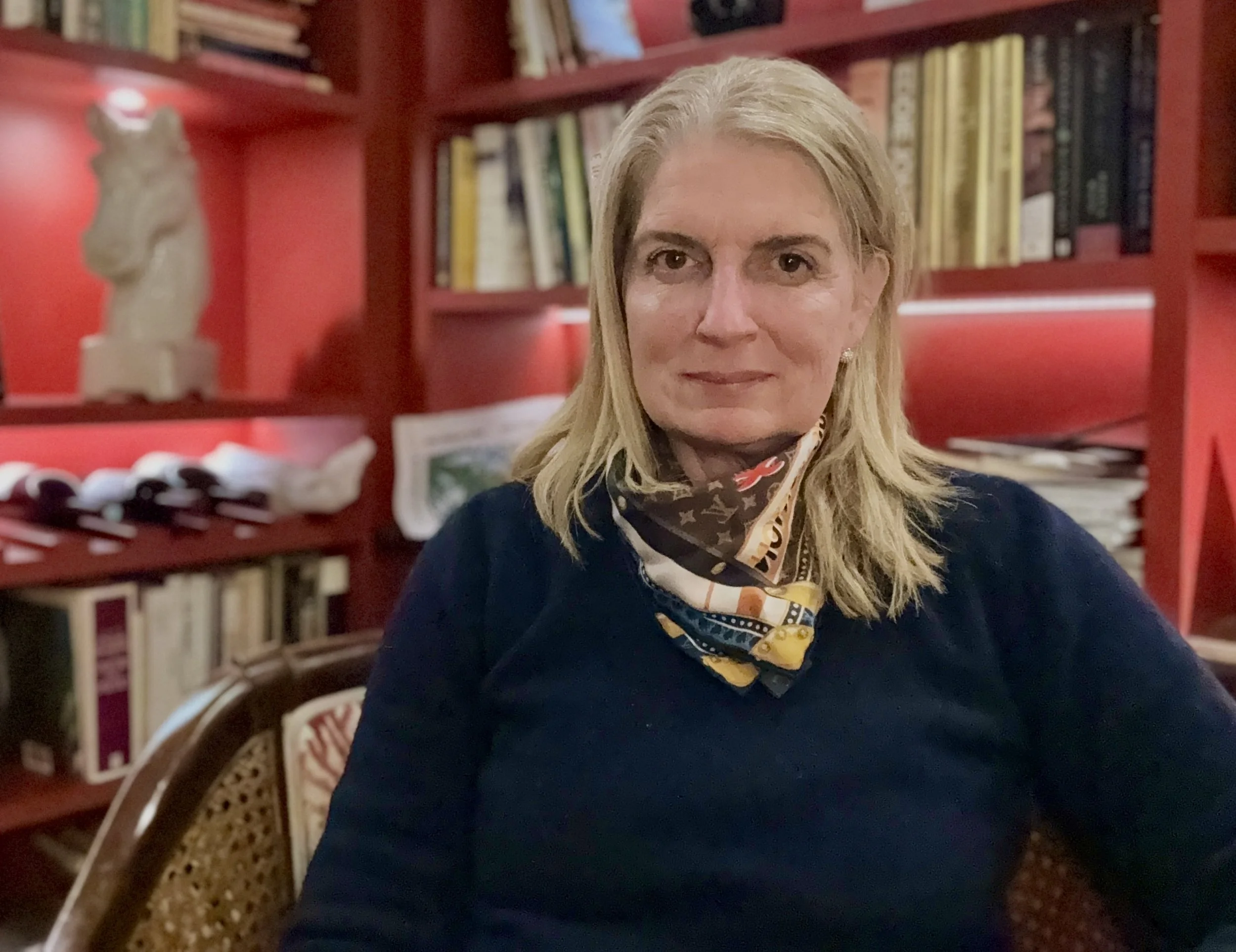 A woman with blonde hair sitting indoors in front of a red bookshelf filled with books and decorative items, wearing a black top and a patterned scarf.
