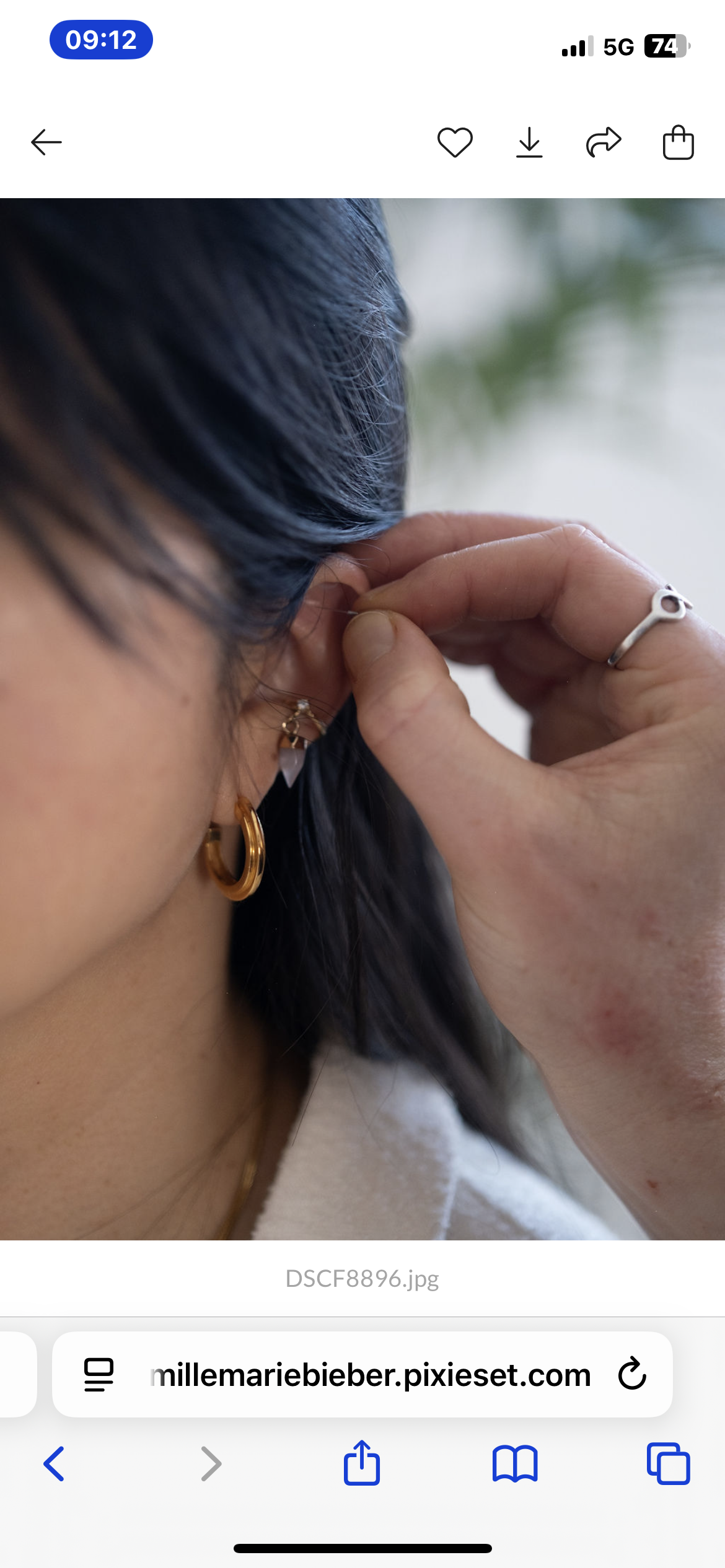 Close-up of a woman putting on an earring, with dark hair, a gold hoop earring, a silver ring on her finger, and a white outfit.