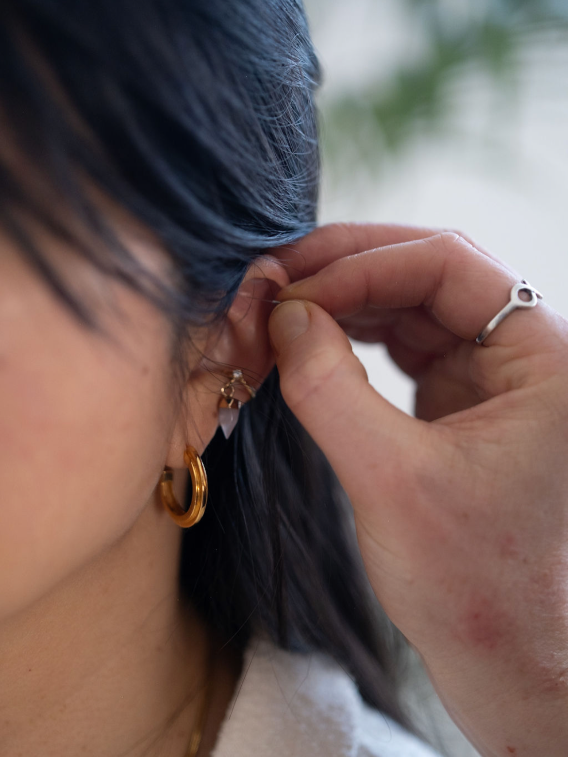A person with dark hair wearing gold hoop and white earrings, inserting earrings into her ear