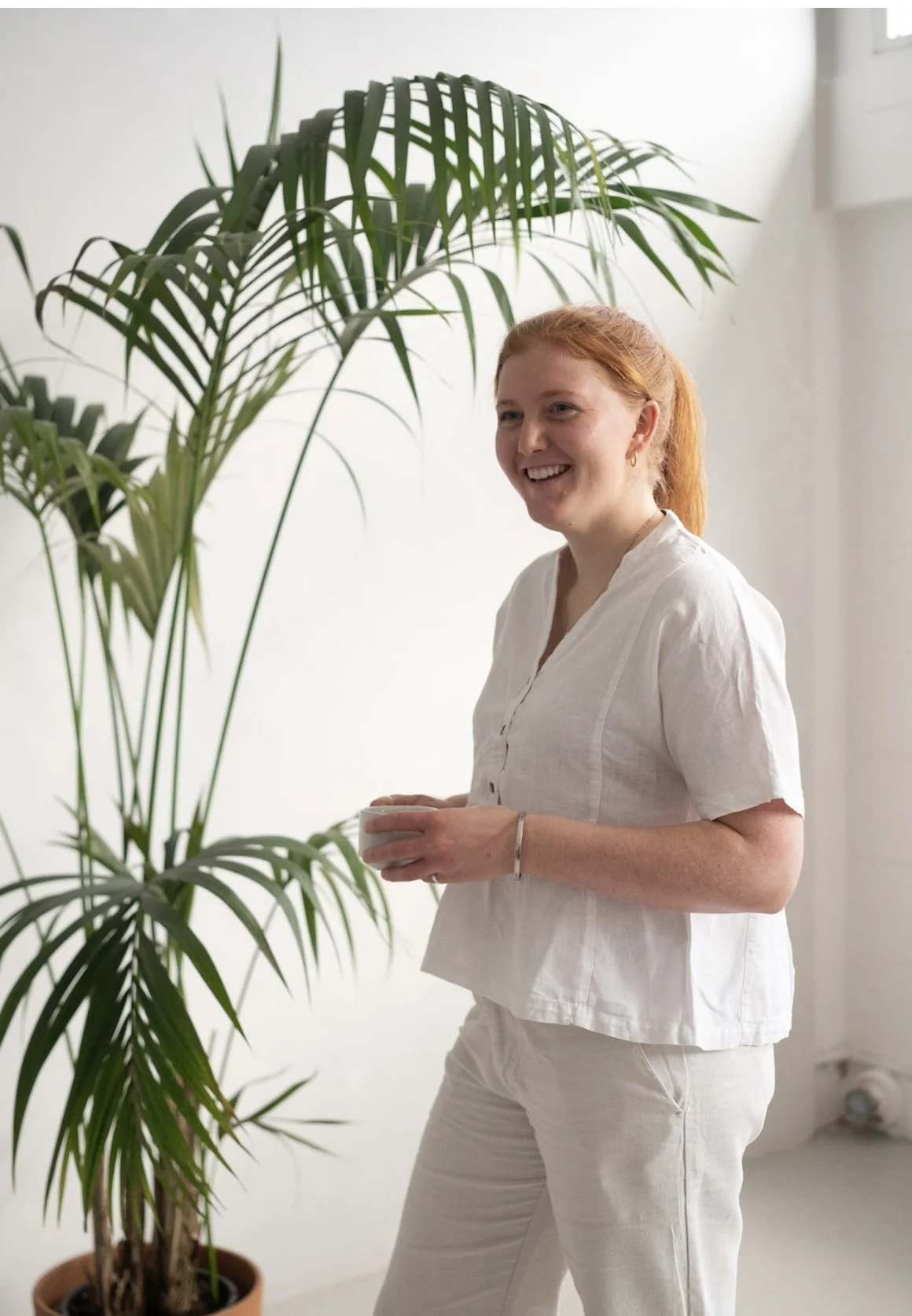 A woman with red hair smiling, wearing a white shirt and pants, standing near a large indoor plant in a white room.