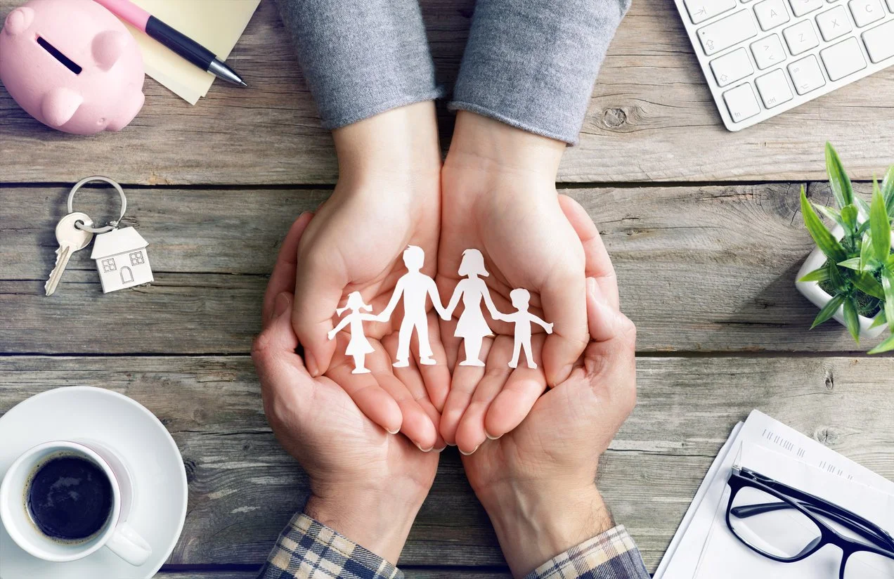 Hands holding paper cutouts of a family of four on a wooden desk with office items around, including a piggy bank, keys, a coffee cup, glasses, a plant, and a keyboard.