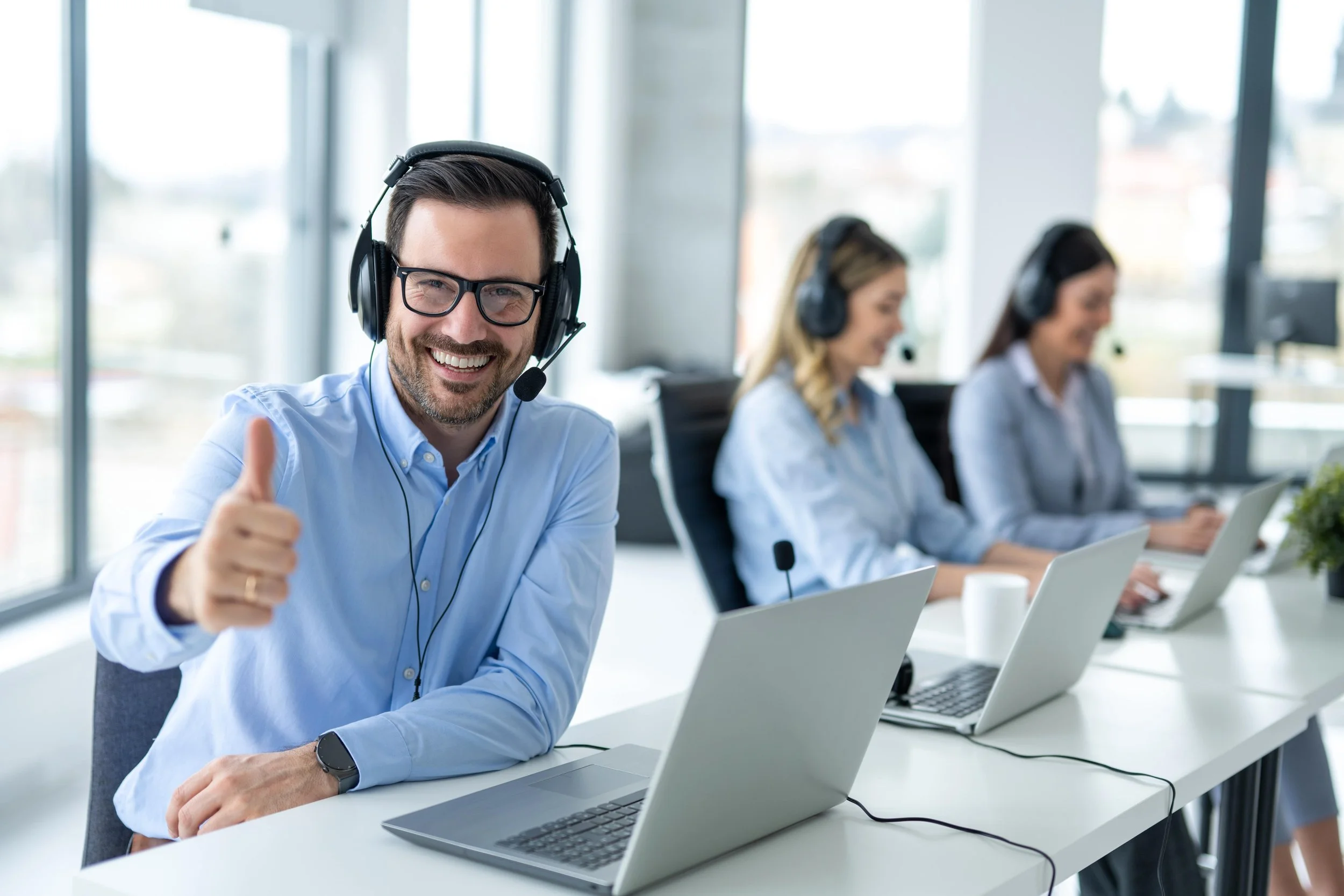 Call center agents working at desks with laptops, wearing headsets, one smiling and giving a thumbs-up.