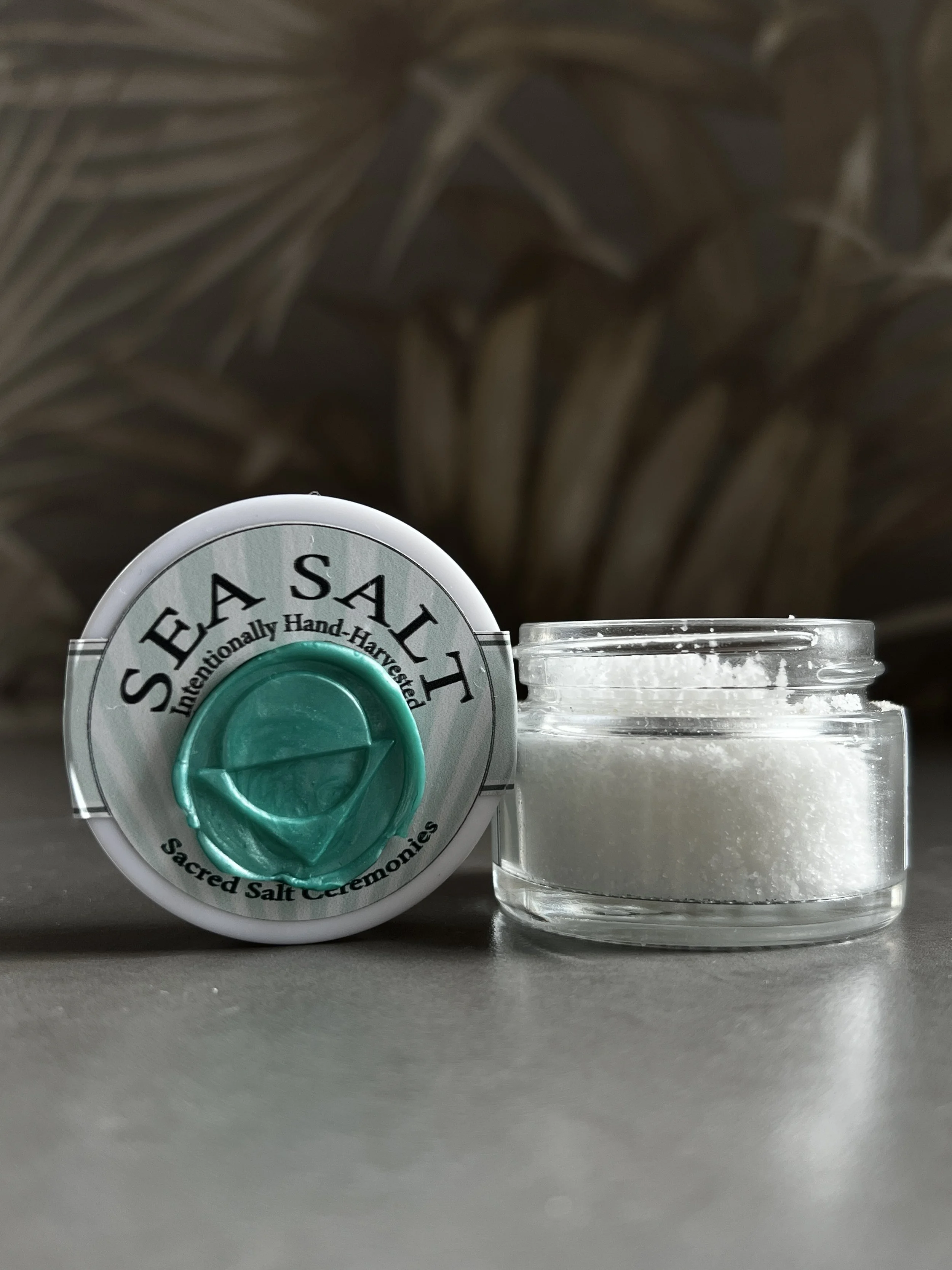 A container of Sacred Salt Ceremonies with a teal and gray label, next to a small glass bowl filled with white salt, with a blurred background of abstract leaves or plant pattern.