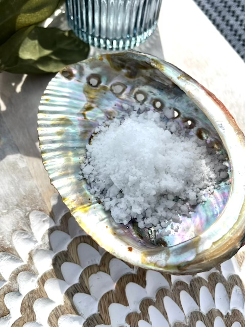 A shell-shaped dish filled with coarse sea salt, placed on a ceremonial table with a glass and greenery nearby.