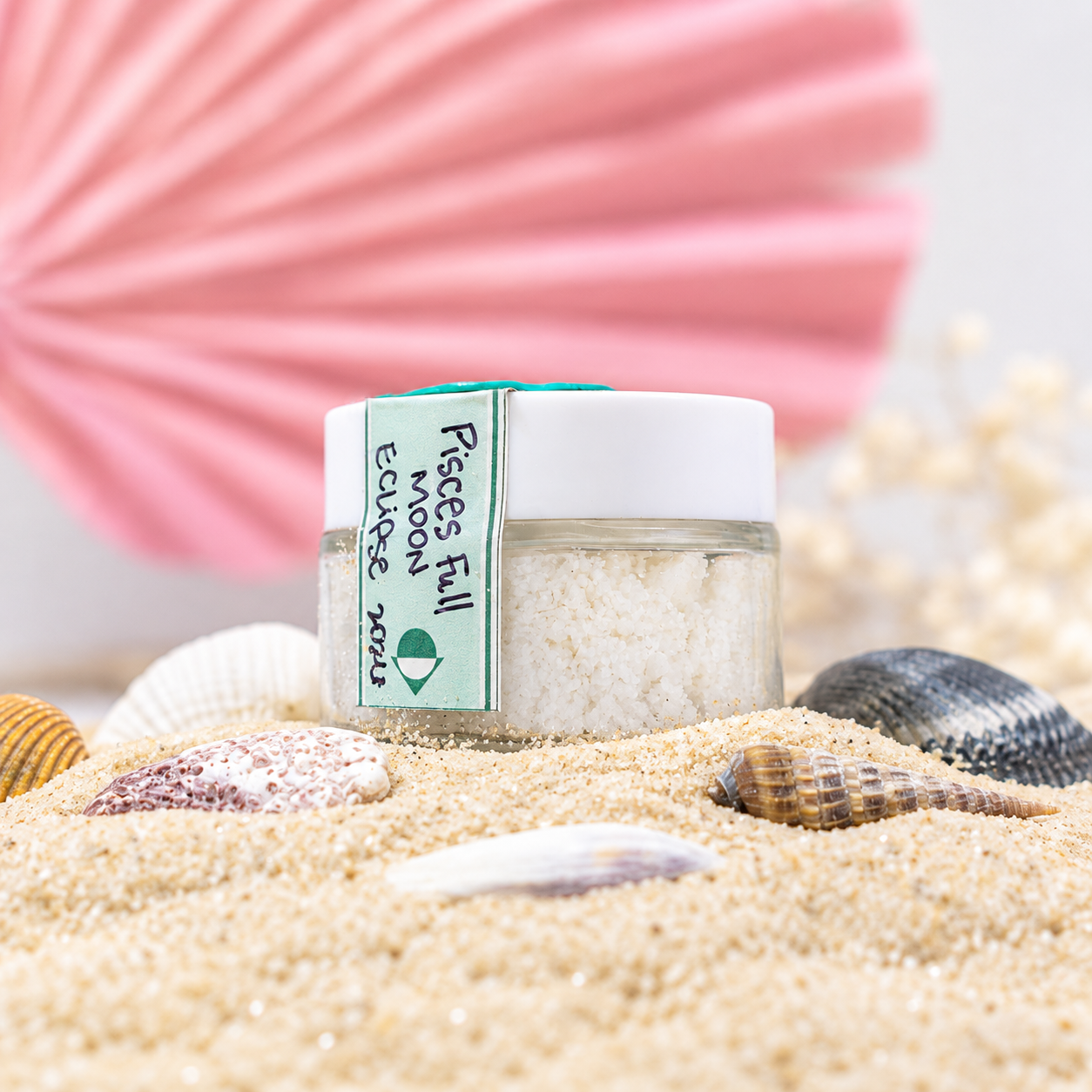 Small jar of bath salts labeled 'Piscees Full Moon December 2023' on sandy surface with seashells and a pink paper fan in background.