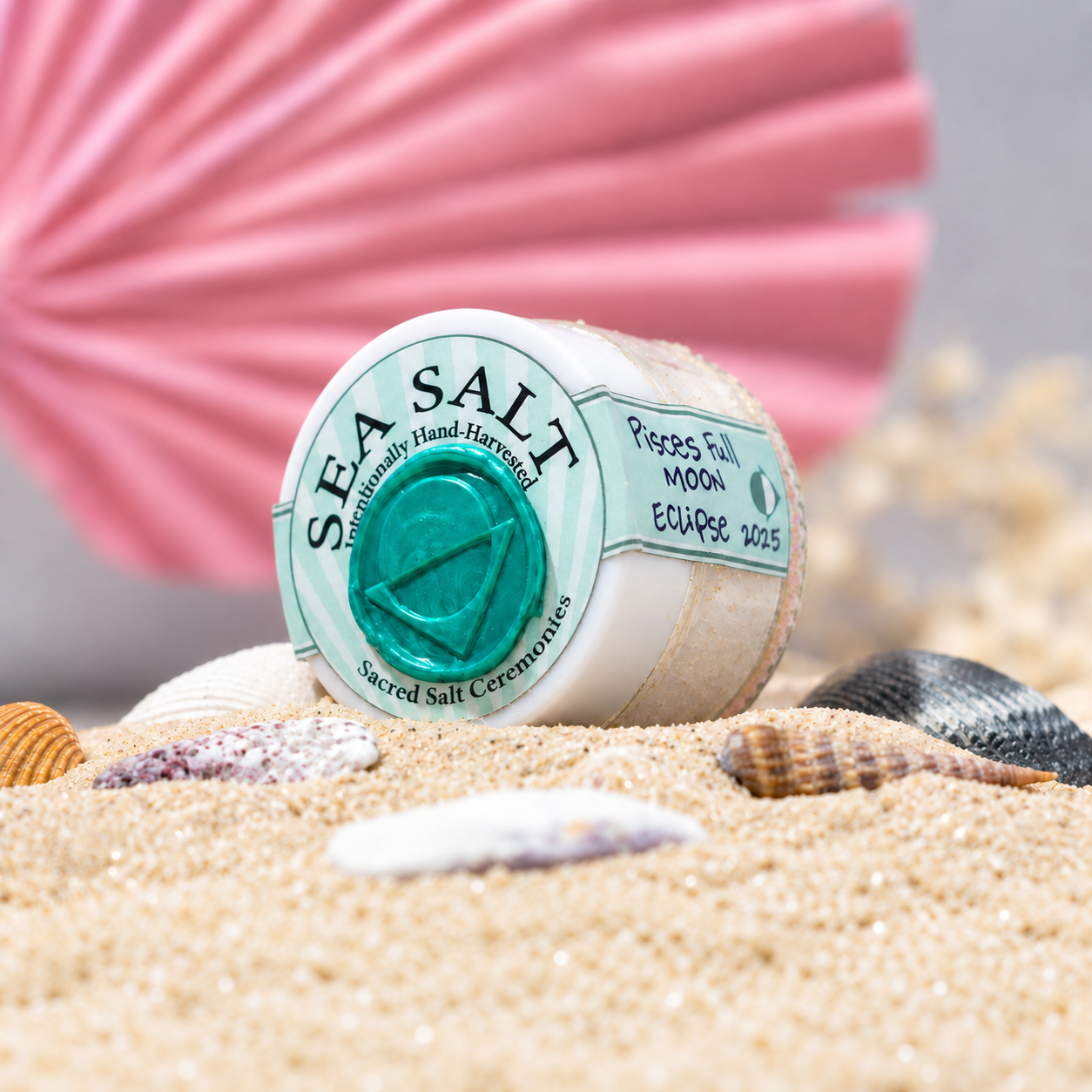 A container of sacred salt from a sacred salt ceremony with star signs, placed on sand along with seashells and with a pink seashell and small white coral in the background.