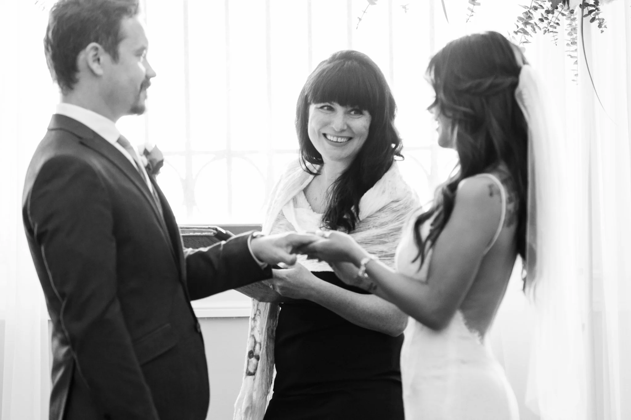 A black and white photo of a wedding ceremony with a couple exchanging rings, a priest or officiant smiling, and an indoor setting with a window in the background.