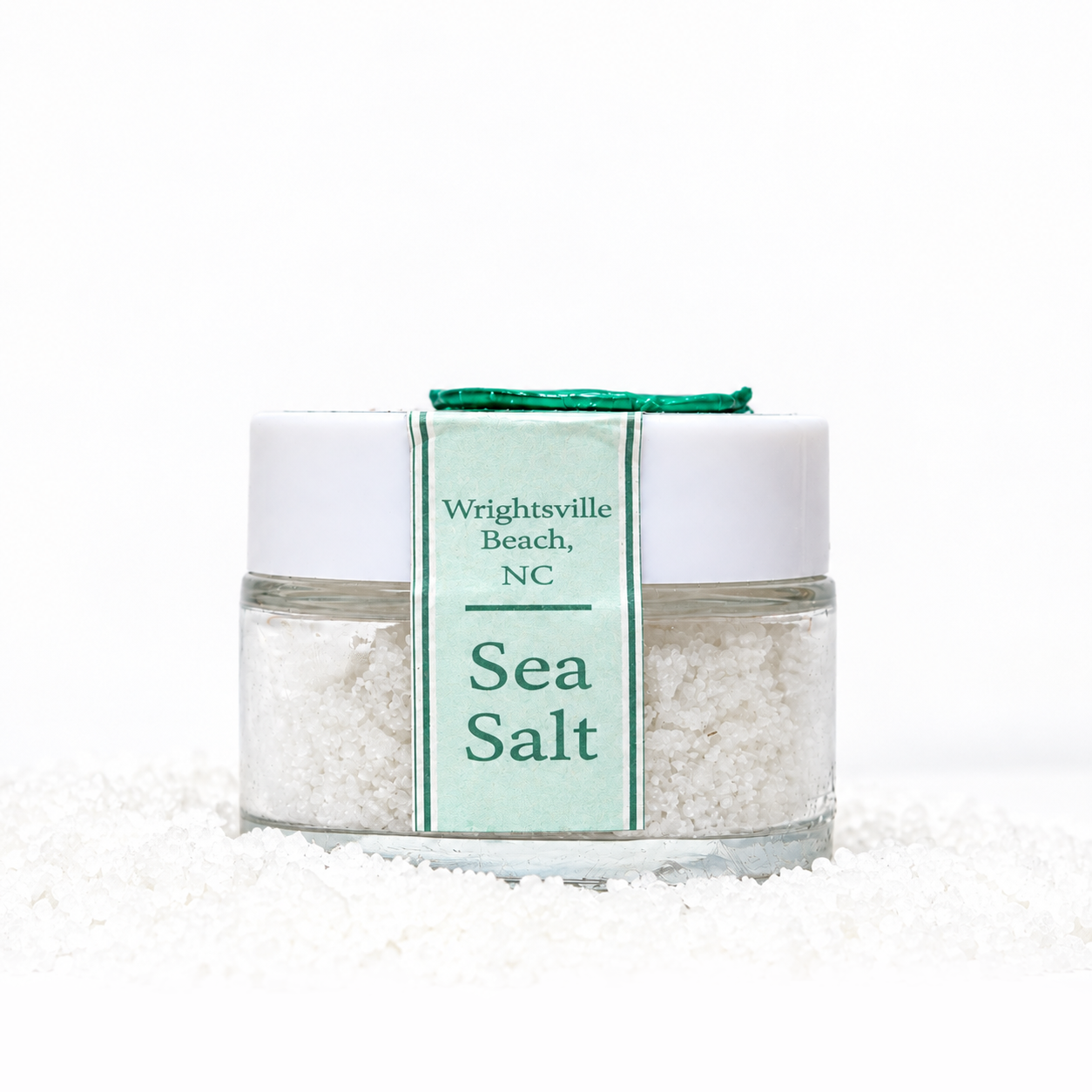 A clear jar of sea salt with a white lid, placed on a surface with spilled salt, and a green label reading 'Wrightsville Beach, NC Sea Salt' sticker sealing the lid.
