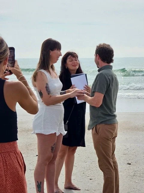 A couple is on the beach during a wedding ceremony, with a woman officiating. A woman is taking a photo with her phone.