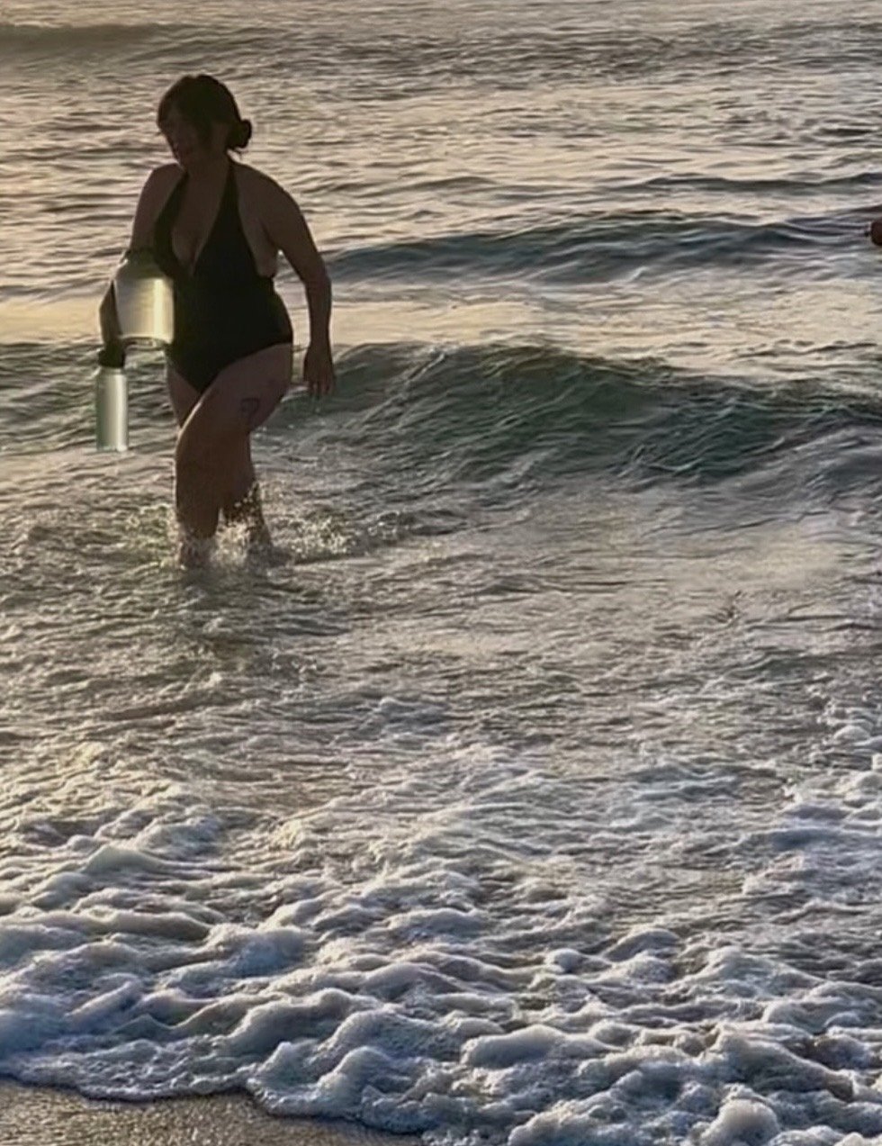 A woman in a black swimsuit walking in the ocean at sunset, carrying a jar of Wrightsville Beach water to distill into sacred salt.