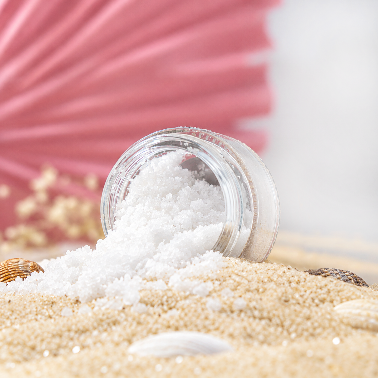 Open container of white sea salt spilling onto sandy surface, with seashells and a pink fan in the background.