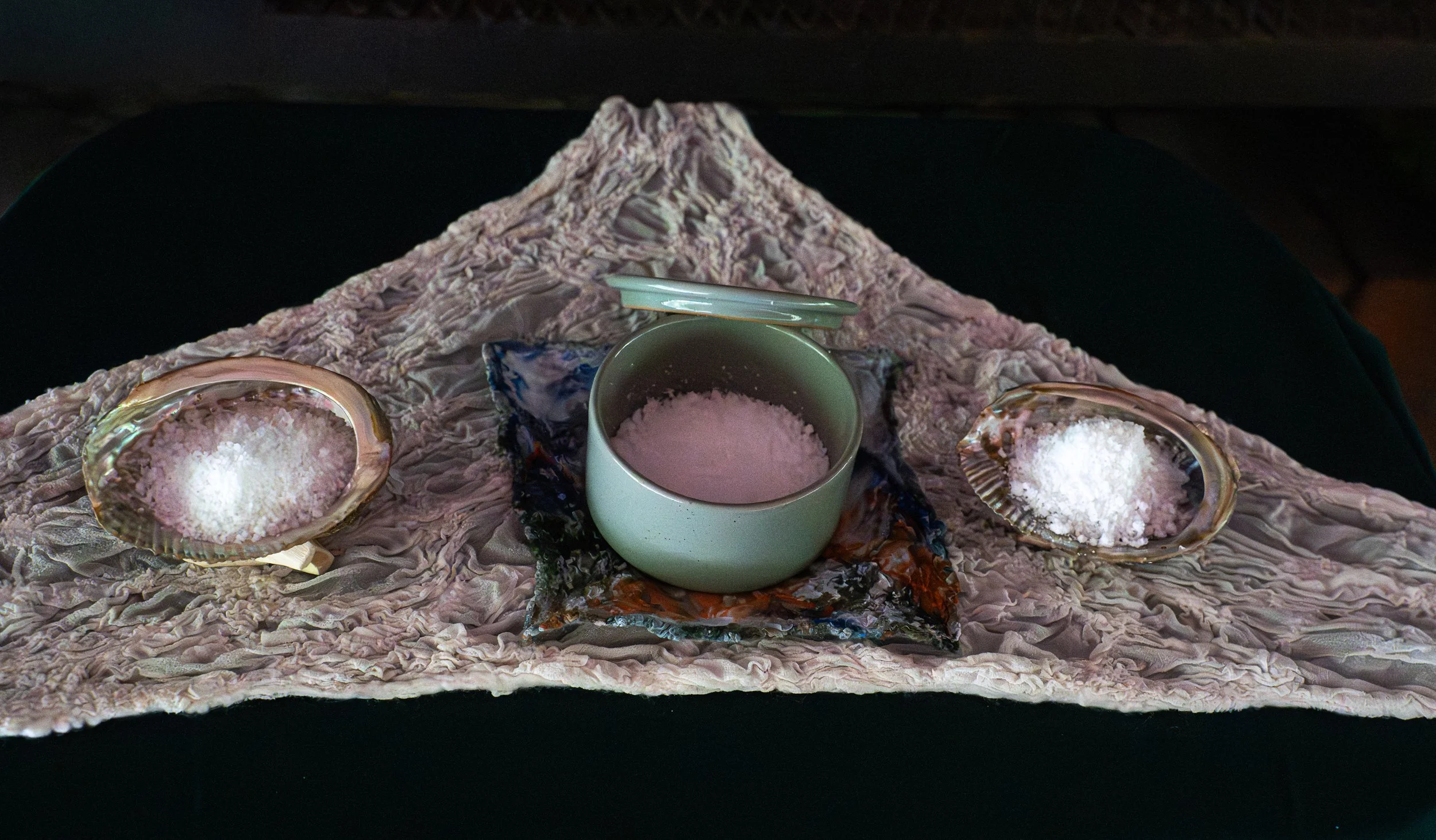 A layered art piece resembling Mount Fuji with a central cup filled with pink powder and two shells filled with white material on either side, all placed on a textured surface.