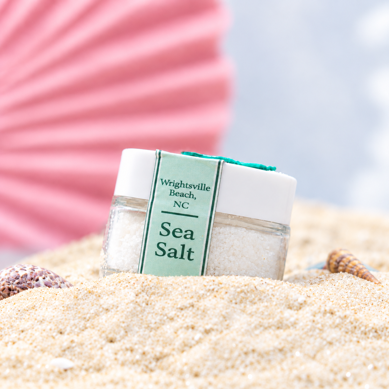Container of sea salt on a sandy beach with seashells and a pink paper fan in the background