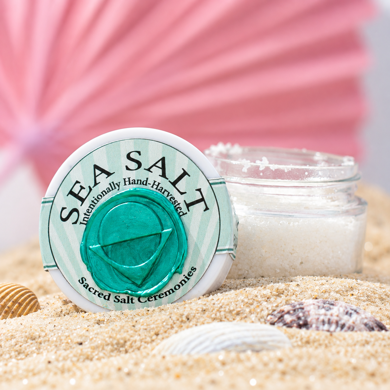 A container of sea salt with a label that reads "Sea Salt, Internationally Hand-Harvested, Sacred Salt Ceremonies" and some seashells resting on sand in the foreground, with a blurred pink seashell in the background.